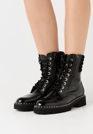 Black patent leather ankle boots with front laces, side zippers, studded trim on thick soles, worn by a person with bare legs.