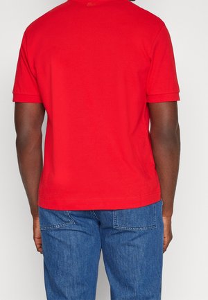 Homme portant un polo rouge à manches courtes et un jean bleu, vu de dos sur un fond uni.