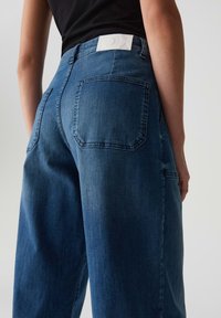 Blauwe denim jeans met een hoge taille en wijde pijpen. Bevat een wit logo label op de achterkant en twee achterzakken met gestikte details.