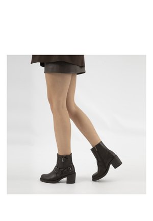 Donna che indossa stivaletti in pelle nera con fibbie e tacchi a blocco, abbinati a una gonna nera, che cammina su uno sfondo bianco uniforme.