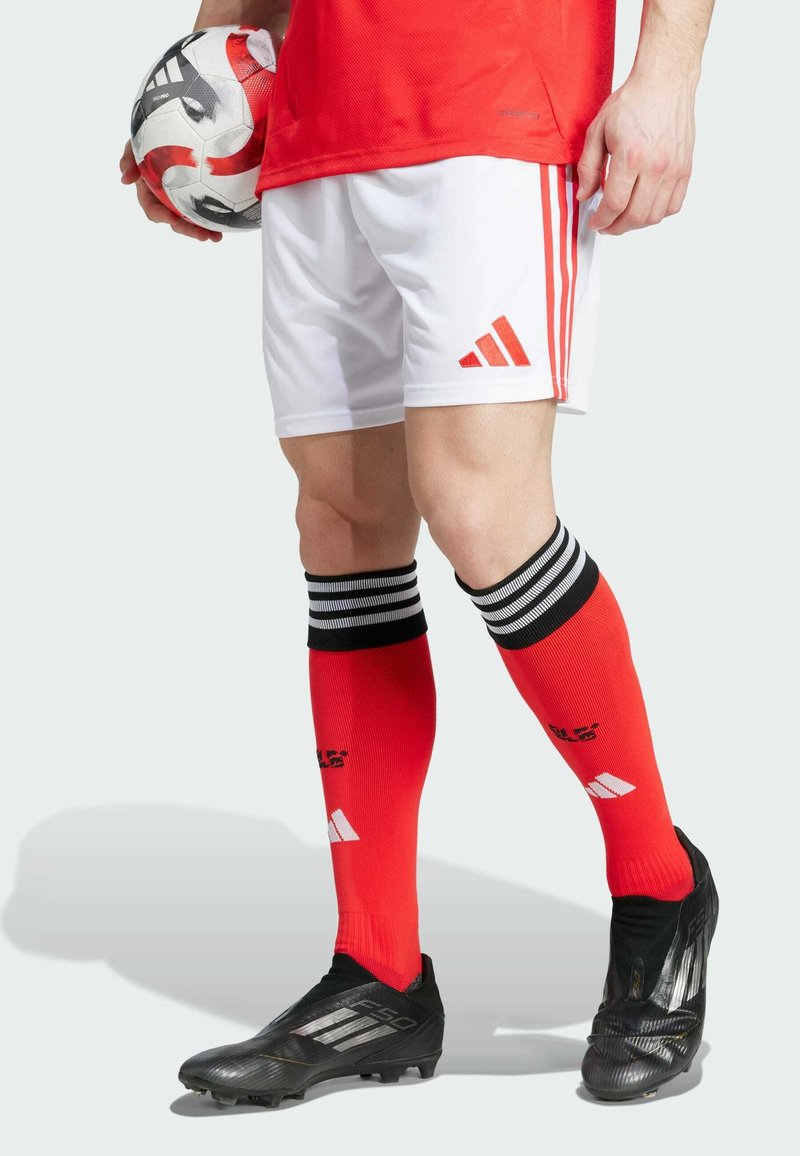 Rotes Sporttrikot, weiße Shorts mit roten Streifen, rote Socken mit schwarzen Streifen, schwarze Fußballschuhe, hält einen schwarz-weißen Fußball.