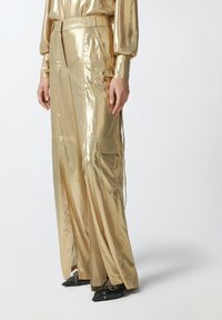 Metalne zlatne široke hlače s sjajnom teksturom, s bočnim džepovima i dizajnom s pojasom. Crne cipele s oštrim vrhom i zakovicama.