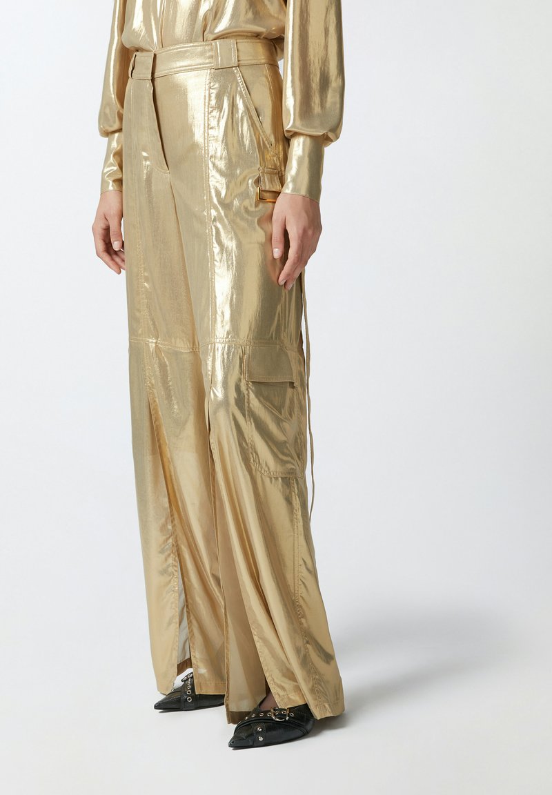 Metalne zlatne široke hlače s sjajnom teksturom, s bočnim džepovima i dizajnom s pojasom. Crne cipele s oštrim vrhom i zakovicama.