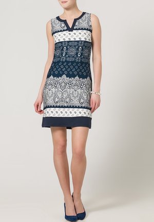 Žena v bezrukávových námořnických a bílých šatech po kolena s propracovanými květinovými a paisley vzory, doplněná námořnickými lodičkami a náramkem.