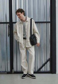 Světle béžová denimová bunda s předními kapsami, šedá košile pod ní, odpovídající béžové kalhoty a černé tenisky. Taška přes rameno visí na rameni.