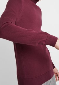 Maglione a collo alto in maglia bordeaux con un design aderente, polsini a coste e una superficie liscia. Presenta dettagli sui lati.