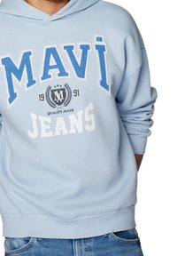 Jasnoniebieska bluza z kapturem z napisem „MAVI JEANS” oraz logo w kolorach niebieskim i białym, noszona z niebieskimi dżinsami, częściowo widoczny tułów i ramiona.