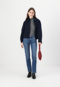 Giacca corta blu navy con collo, texture a maglia e chiusura con zip, abbinata a una maglietta grigia e jeans blu. Stivali neri alla caviglia e borsa rossa.
