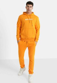 Sudadera naranja con bolsillo frontal y cordón, con un logo blanco. Combinada con joggers naranjas a juego y zapatillas blancas.
