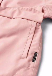 Giacca rosa chiaro con una texture liscia, tasca laterale e un'etichetta nera rettangolare con un motivo a griglia in basso a sinistra.