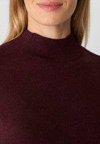 Donna che indossa un maglione aderente a coste bordeaux con collo alto, mostrando il collo, le spalle e la parte bassa del viso.