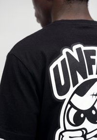 Person trägt ein schwarzes T-Shirt mit großem weißem "UNFC"-Grafik und einer Cartoonfigur auf dem Rücken.
