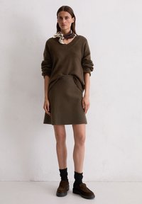 Vrouw die een bruine V-hals trui en bijpassende rok draagt, donkere sokken, bruine loafers en een bruin-crème sjaaltje geknoopt rond haar nek.