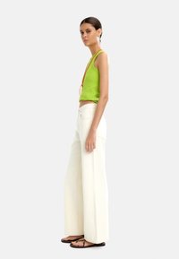 Zielony prążkowany top na ramiączkach zestawiony z białymi spodniami typu wide-leg o wysokim stanie. Modelka nosi brązowe klapki i naszyjnik z zawieszką.