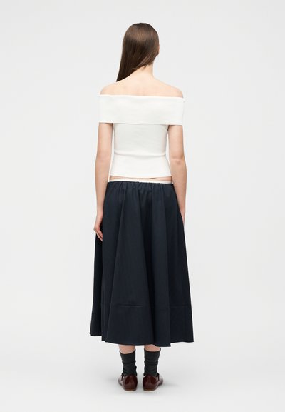 Femme aux longs cheveux bruns portant un haut blanc à épaules dénudées, une jupe midi noire, des chaussettes foncées et des chaussures marron, debout de dos.