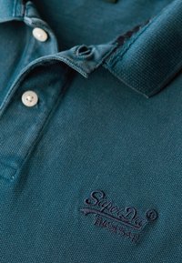 Polo de color verde oscuro con tejido texturizado, dos botones blancos y logo bordado "SuperDry" en azul marino. Diseño con cuello y abundante costura.