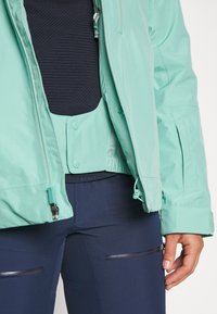 Helle teal wasserdichte Jacke mit Reißverschluss und Kordelzug in der Taille, über einem strukturierten navyfarbenen Oberteil und navyfarbenen Hosen mit Reißverschlusstaschen getragen.