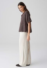 Blusa marrone strutturata con texture plissettata, maniche corte, abbinata a pantaloni bianchi a gamba larga e scarpe nere in tessuto a slip-on.