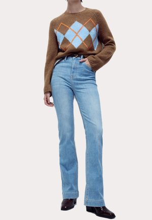 Osoba ubrana w brązowy sweter z niebiesko-pomarańczowym wzorem diamentowym, jasnoniebieskie, wysokowalne dżinsy oraz ciemnobrązowe buty, stojąca z jedną ręką w kieszeni.