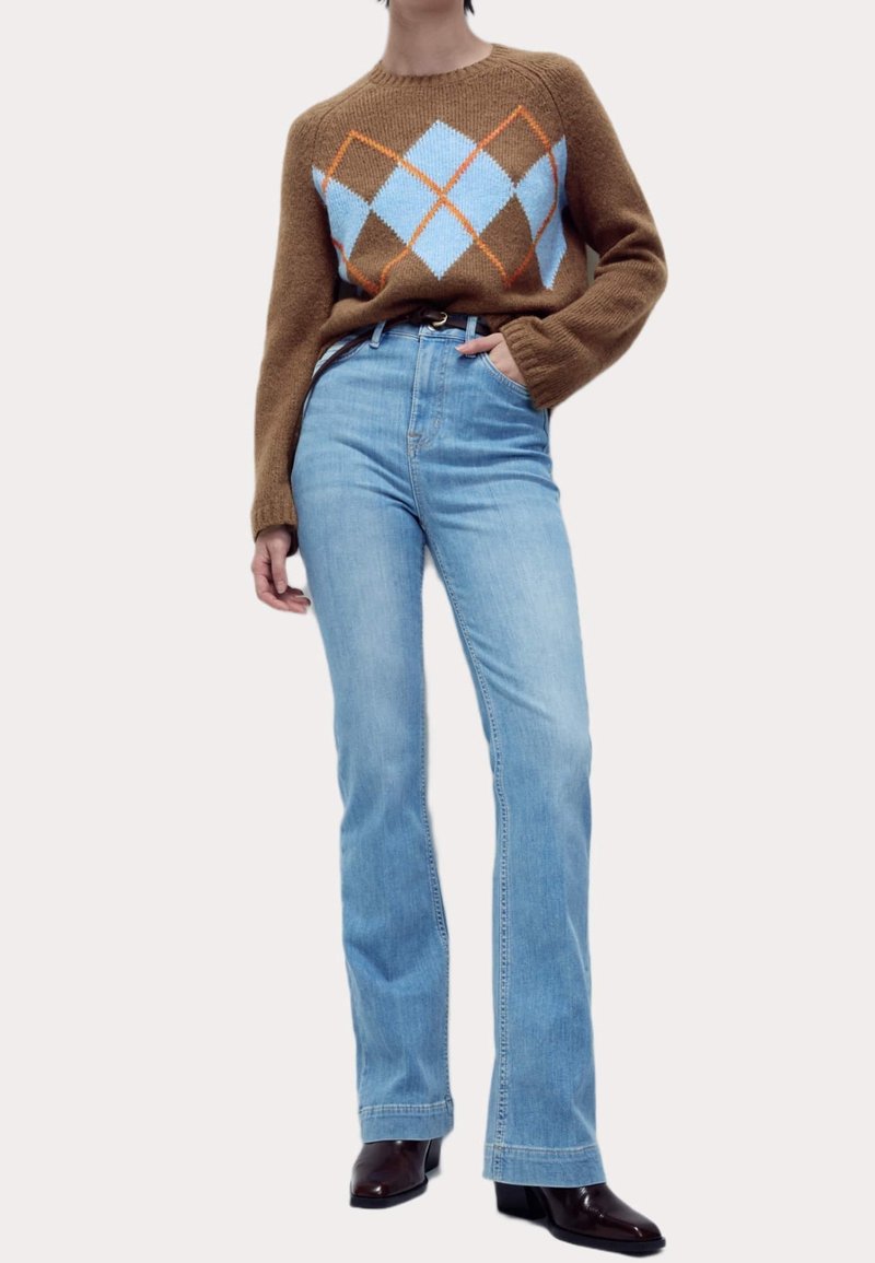 Personne portant un pull marron avec un motif de losanges bleu et orange, un jean taille haute bleu clair et des bottes marron foncé, debout avec une main dans la poche.