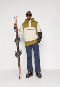 Ένδυση σκι που περιλαμβάνει ένα μπερδεμένο, ελαιόχρωμο και μπεζ μπουφάν με κουκούλα, μπροστινή τσέπη και μαύρες λεπτομέρειες, σε συνδυασμό με μπλε παντελόνι σκι και εξοπλισμό.