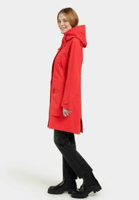Parka imperméable rouge avec capuche, manches longues, poches latérales et coupe droite. Assortie à un jean noir à jambe droite et des bottes noires imposantes.