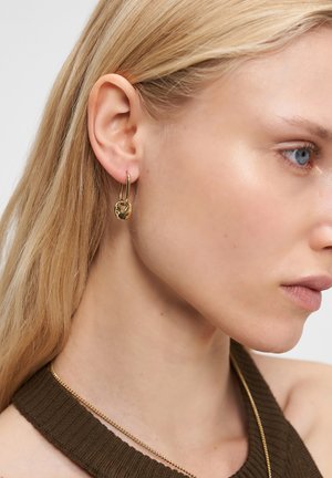 Close-up of a woman with blonde hair wearing gold hoop earrings with a small pendant and a gold chain necklace, dressed in a brown top.