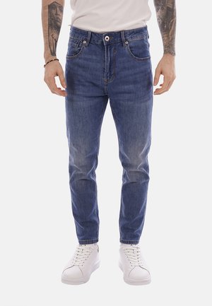 Uomo che indossa jeans blu medio a taglio aderente con tasche anteriori e scarpe da ginnastica bianche, in piedi con le mani lungo i fianchi su sfondo bianco.