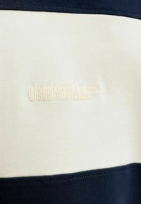 Gros plan sur un tissu crème et bleu marine avec le mot « ORIGINALS » brodé en fil crème sur la partie plus claire.