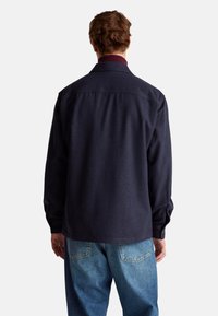 Camisa de botón azul marino oscuro con un cuello burdeos; presenta mangas largas y un corte relajado. Se lleva con jeans de mezclilla azul claro.