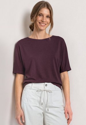 Jeune femme souriante portant un haut violet à manches courtes et un pantalon blanc avec une taille à cordon, devant un mur clair.