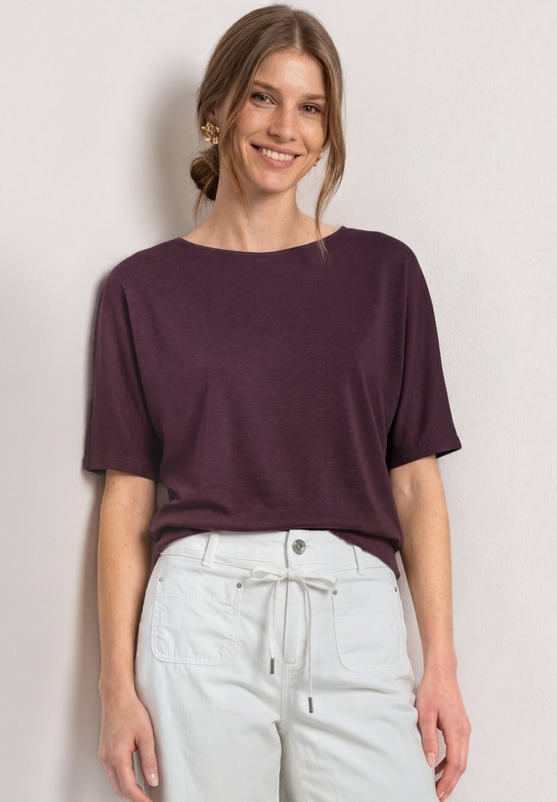Jeune femme souriante portant un haut violet à manches courtes et un pantalon blanc avec une taille à cordon, devant un mur clair.