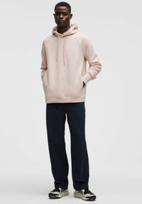 Sudadera rosa clara con un bolsillo tipo canguro, puños acanalados y cordones, combinada con pantalones azul marino oscuros y zapatillas negras y blancas.
