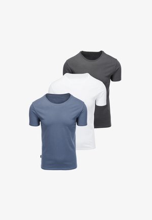 Mann mit Bart, der ein eng anliegendes graues T-Shirt mit kurzen Ärmeln, blaue Jeans und eine schwarze Armbanduhr vor einem schlichten hellen Hintergrund trägt.