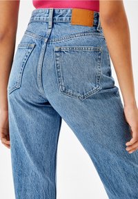 Person som bär högmidjade ljusblå denimjeans med bakfickor och en brun läderloggopatch, i kombination med en röd topp.