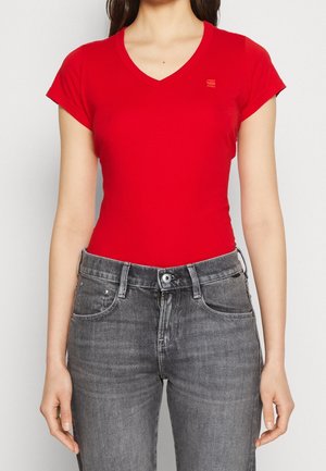Parte superior del cuerpo de una persona que lleva una camiseta ajustada de manga corta con cuello en V de color rojo, combinada con jeans de mezclilla gris de cintura alta, sobre un fondo blanco liso.