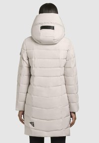 Padded coat in light beige with a hood. It features a quilted design, a black logo patch on the back, and a smooth texture.
