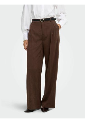 Personne portant un pantalon marron taille haute à jambes larges avec plis, ceinture noire, chemise blanche à manches longues et chaussures noires à bouts pointus, debout avec une main dans la poche.