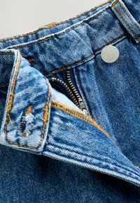 Jean en denim avec une fermeture éclair, des coutures en cuivre et un bouton argenté. Le tissu est d'un bleu clair avec une texture douce.