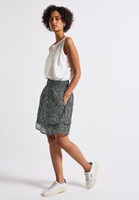 White sleeveless top with textured straps, paired with a green skirt featuring a leaf pattern, side pockets, and white sneakers.