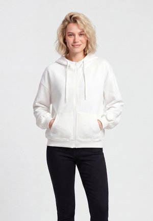 Persoană blondă purtând un hanorac alb cu fermoar, având mâinile în buzunare și pantaloni negri, stând în fața unui fundal simplu și deschis.