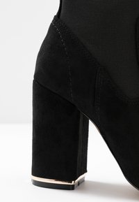 Stivale nero con tacco alto, dalla superficie in suede elegante, tacco quadrato e un sottile accento dorato alla base del tacco. Dettaglio in tessuto liscio sopra.