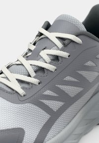 Gray athletic shoe with a textured mesh upper, rubber sole, and white laces. Features a smooth overlay and angled patterns on the side.