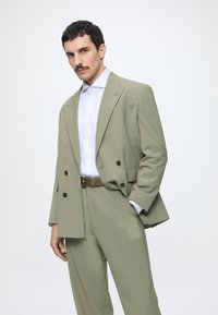 Homme avec moustache portant un costume croisé olive clair, chemise blanche et ceinture marron, debout avec une main dans la poche devant un fond uni.