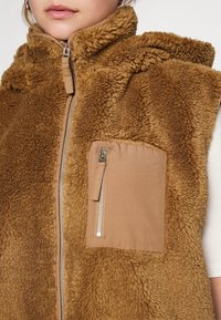 Braune Kunstfell-Weste mit hohem Kragen, ausgestattet mit einem Front-Reißverschluss und einer beigefarbenen Stofftasche mit einem Reißverschluss auf der linken Brust.