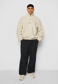 Kremfarget, fuzzy fleece genser med høy krage og halv glidelås, kombinert med løse svarte cargo bukser; begge har broderte logoer.