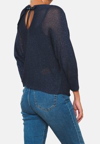 Navy Pullover mit strukturiertem Strick, offener Rückseite und Ripsband-Detail. Kombiniert mit eng anliegenden blauen Jeans mit Gesäßtaschen.