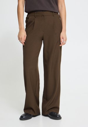 Persona con pantalones de vestir marrones holgados y zapatos negros, de pie contra un fondo liso y claro. Solo son visibles el torso inferior y las piernas.