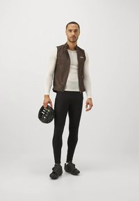 Mann iført en brun vest, hvit langermet skjorte, svarte leggings og sko, som holder en svart sykkelhjelm mot en ensfarget bakgrunn.