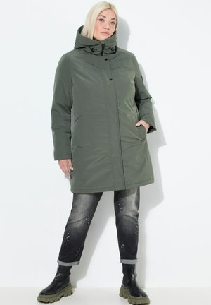 Vrouw met blond haar, gekleed in een olijfgroene gewatteerde jas met capuchon, zwart-grijze opgerolde jeans met versieringen en zwarte chunky laarzen.